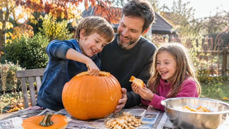 Vater mit kurzem Bart und zwei lachende Kinder schnitzen im Herbstgarten einen großen orangefarbenen Kürbis auf zeitungsbedecktem Tisch