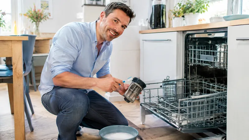Lächelnder Mann in hellblauem Hemd hockt vor einer geöffneten Spülmaschine, hält das herausgenommene Sieb und eine alte Zahnbürste in den Händen, davor eine Schüssel mit Spülwasser